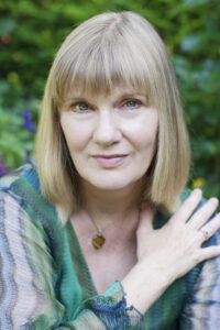 About me, Laurelle Rond. Laurelle has shoulder length blonde hair and a blunt fringe, she is sitting in front of green nature and has one hand gently rested on her shoulder.
